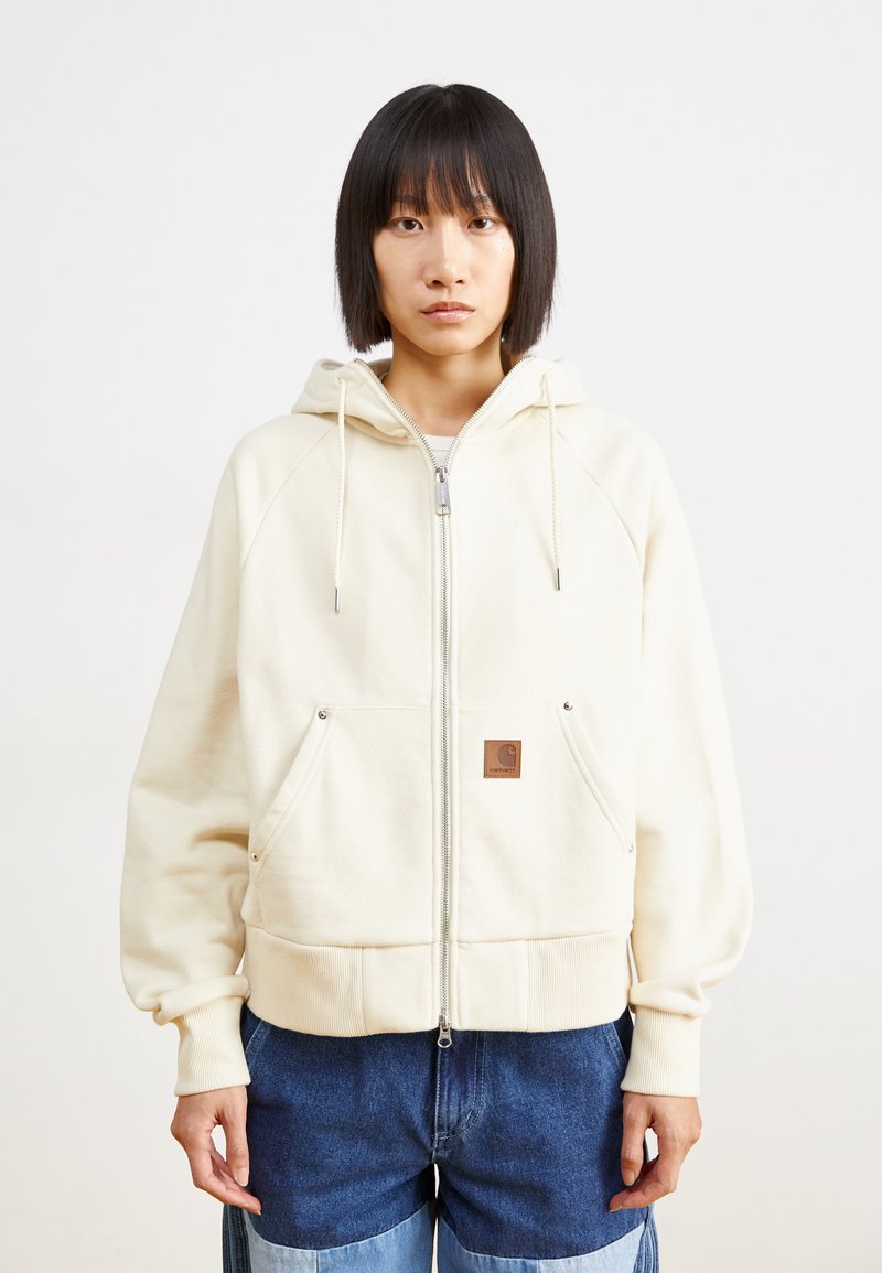 Hoodie zippé crème avec une capuche à cordon, des poches avant et un écusson de logo marron, ayant une coupe décontractée et des poignets et ourlet en ribbed.