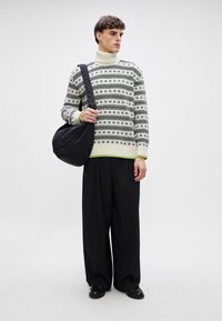 Strikket genser i svart og krem med geometriske mønstre, kombinert med løse svarte bukser og en svart quiltet skulderveske.