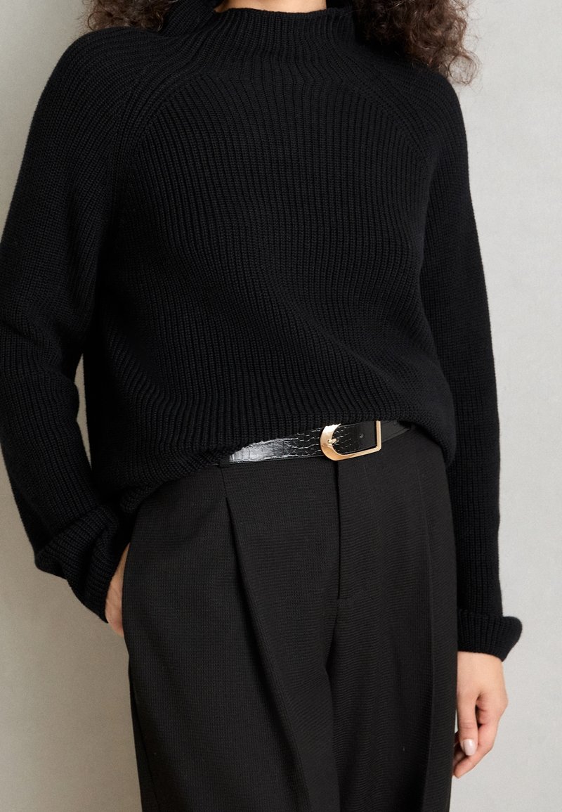 Maglione nero a maglia con collo alto, texture a costine e maniche lunghe, abbinato a pantaloni neri a vita alta e una cintura nera lucida.