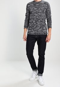 Maglione a maniche lunghe con scollo rotondo in un motivo mélange bianco e nero, abbinato a jeans in denim scuri e sneakers bianche con cinturini visibili.