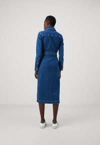 Denimklänning, långärmad, längd till mitten av vaden, bälte i midjan, blåtvätt, knäppta manschetter, enkel krage, bärs med vita sneakers.