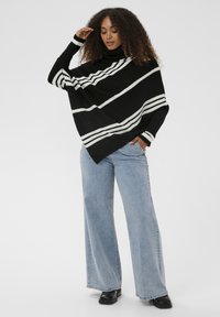 Moteris, vilkinti juodai-baltą dryžuotą megztinį su aukštu kaklu, šviesiai mėlynus plačius džinsus ir juodus batus, pozuojanti su ranka liečiančia plaukus.