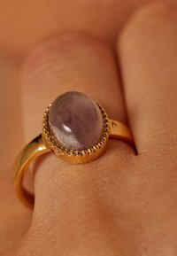Gouden ring met een ovale, lichtpaarse steen. De band heeft een gladde afwerking met een kralenstructuur rond de rand van de steen.