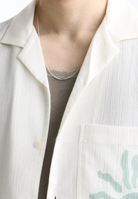 Persona vistiendo una camiseta gris acanalada debajo de una camisa blanca texturizada con un bolsillo que presenta un diseño abstracto en verde claro y un collar de cadena de plata.