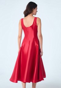 Robe en satin rouge mi-longue avec une jupe évasée, un dos plongeant et de larges bretelles. Texture lisse, design minimaliste sans motifs ni ornements.