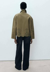 Veste vert olive avec un col haut et un ourlet festonné, présentant des motifs floraux découpés. Associée à un pantalon noir à jambes larges.
