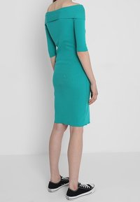 Türkisfarbenes Off-Shoulder-Kleid mit kurzen Ärmeln, figurbetontem Design, knielangem Saum, kombiniert mit schwarzen Sneakers. Glattes, dehnbares Material.