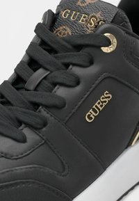 Baskets noires Guess avec logo doré sur le côté et la languette, lacets noirs et détail texturé à motif sur la languette.