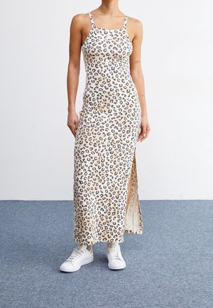 Vrouw draagt een beige leopardprint mouwloze maxi-jurk met zijsplit en witte sneakers, staand op een blauw tapijt.