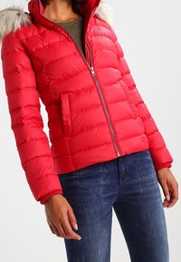 Czerwona kurtka puchowa z zamkiem z przodu i kapturem z sztucznego futra. Posiada pikowaną konstrukcję, ściągacze przy mankietach oraz boczne kieszenie. Nosi się ją na zielonej bluzce.