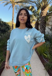 Felpa azzurro chiaro con grafica di cuore e occhio, vestibilità comoda e ricamo "HOPE". Abbinata a leggings colorati con motivi.