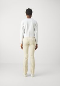 Vit långärmad topp med tryck på baksidan, kombinerad med beiga slim-fit-byxor. Modellen bär vita skor. Minimalistisk design med mjuk textur.