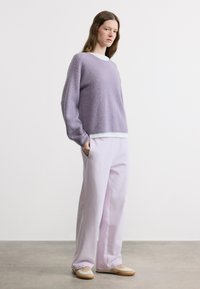 Γυναίκα που φοράει ανοιχτό μοβ πλεκτό πουλόβερ, ταιριαστό παντελόνι με φαρδιά πόδια, μπεζ αθλητικά παπούτσια, στέκεται με το ένα χέρι στην τσέπη πάνω σε γκρι πάτωμα.