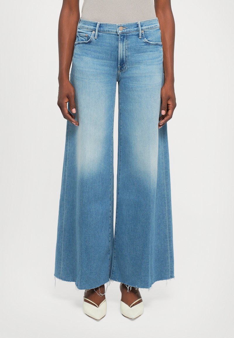 Person wearing light blue wide-leg jeans with raw hems and white pointed-toe heels against a plain background.