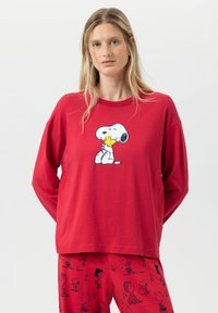Een vrouw met lang blond haar draagt rode Peanuts-thema pyjama's met Snoopy die Woodstock omarmt op de bovenkant en Charlie Brown-personages op de broek.