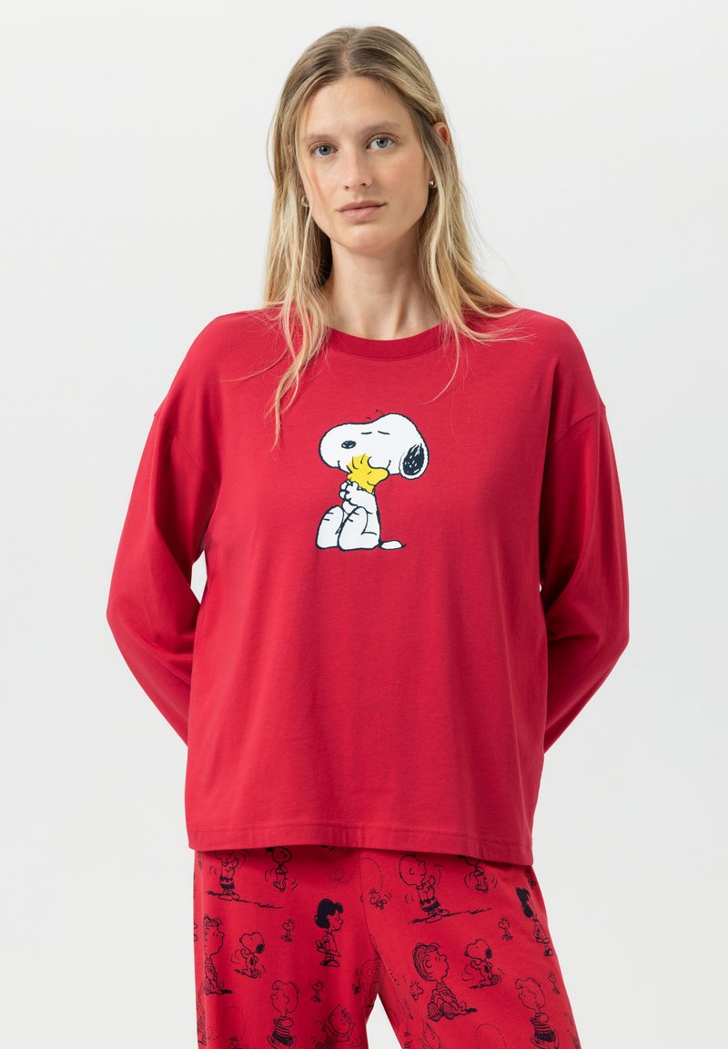 Een vrouw met lang blond haar draagt rode Peanuts-thema pyjama's met Snoopy die Woodstock omarmt op de bovenkant en Charlie Brown-personages op de broek.