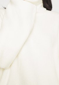 Vit, ribbad polotröja med långa ärmar. Mjuk textur, lös passform, har en hög krage. Inga synliga mönster eller detaljer.