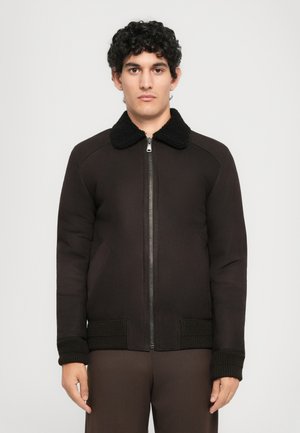 Young man with curly dark hair wearing a dark brown zip-up jacket with a black fleece collar and knitted cuffs, standing against a plain background.