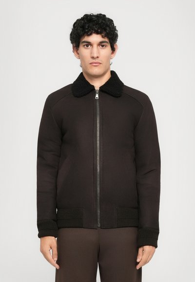 Jonge man met krullend donker haar, gekleed in een donkerbruine zip-up jack met een zwarte fleece kraag en gebreide manchetten, staand tegen een effen achtergrond.