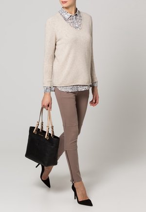 Pull beige avec un col en V sur une chemise à motifs, associé à un legging taupe et des chaussures noires à talons hauts. Sac à main noir avec des bretelles.