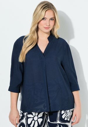 Mujer rubia que lleva una blusa suelta de color azul marino oscuro con mangas tres cuartos y pantalones estampados en blanco y negro, de pie contra un fondo blanco.