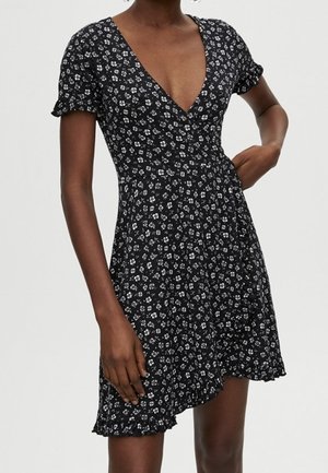 Kvinde iført en kort sort kjole med hvidt blomsterprint, V-udskæring, pufærmer og en rynket asymmetrisk kant.