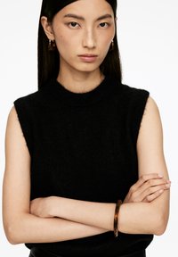 Schwarzes, ärmelloses Wolloberteil mit hohem Ausschnitt. Das Model trägt ein braunes Schildkrötenmuster-Armband am Handgelenk, die Arme verschränkt. Glatte Textur.