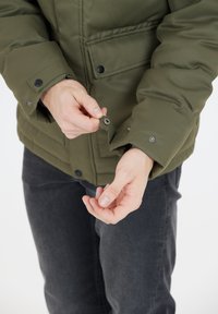 Olivengrønt pufferjakke med knapærme. Har en glat, isoleret tekstur og sidelommer. Mørkegrå jeans synlige nedenfor.