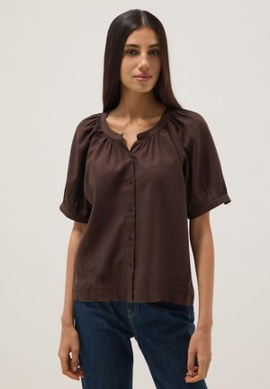 Mujer con cabello largo y oscuro, vestida con una blusa marrón oscuro abotonada con mangas cortas abombadas y vaqueros azules, de pie frente a un fondo liso.