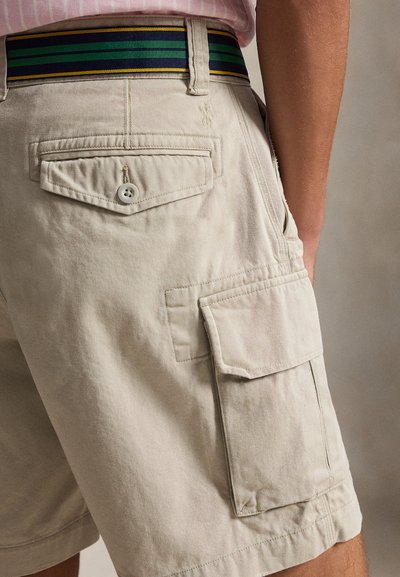 Béžové cargo šortky s gombíkovým zadným vreckom a klopovým bočným vreckom, nosené s pruhovaným opaskom a ružovou košeľou.