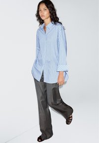 Light blue striped button-up shirt with long sleeves, paired with loose, gray trousers and dark sandals. Minimalist design, smooth fabric.