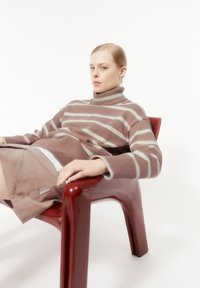 Brunrandig stickad tröja med hög krage, i kombination med en ljusbrun kjol, sittande i en blank röd plaststol mot en vit bakgrund.