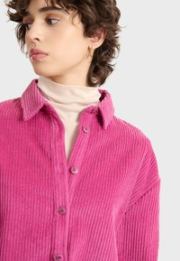 Camisa de pana rosa con cuello, cierre de botones en la parte delantera y textura acanalada. Presenta un corte relajado y detalles de costura visibles.