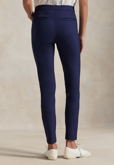 Persoon die strak zittende marineblauwe broek en witte sneakers draagt, met de rug naar de kijker toe staand tegen een effen beige achtergrond.