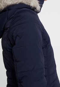Giacca a vento blu navy con cappuccio foderato in pelliccia sintetica, texture imbottita e dettagli di cucitura laterali. Chiusura con zip e tasche visibili di lato.