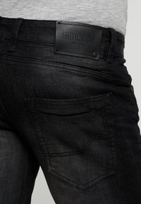 Jean en denim noir avec une poche arrière et une ceinture, patch en cuir noir marqué "INDICODE EST. 1997", porté avec une chemise gris clair.