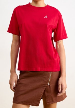 Frau trägt ein rotes T-Shirt mit kurzen Ärmeln und kleinem weißem Logo sowie einen braunen Rock mit Reißverschluss und Schlitz vorne.