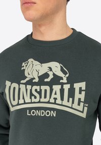 Sötétzöld pulóver, amelyet egy kiemelkedő, textúrált oroszlángrafika és merész "LONSDALE LONDON" felirat díszít világos olívazöld színben.