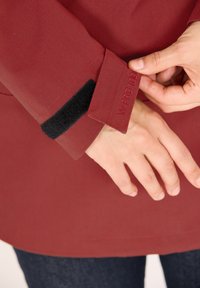 Röd ytterklädnad med en texturerad yta, med en svart kardborrearm och broderad logotyp. Justerar ärmslutet för hand.