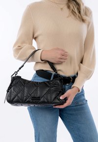 Zwarte gewatteerde handtas met een korte strap, met diagonale stiksels en een soepele textuur, vastgehouden door een persoon in een beige geribbelde trui.