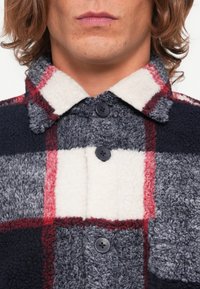 Fleece-Hemdjacket in Marineblau, Grau und Weiß mit Karomuster. Mit einem gemütlichen Kragen und zwei schwarzen Knöpfen an der Vorderseite.