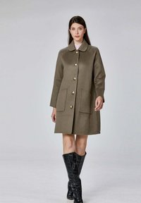 Manteau en laine vert olive avec un col rond, des boutons dorés et deux poches avant. Porté avec des bottes hautes noires brillantes.