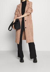 Abrigo largo de tweed en rosa y naranja con un diseño estructurado, que presenta grandes botones negros y una abertura en la parte inferior, llevado sobre pantalones negros y botas.