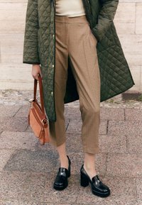 Cappotto imbottito verde oliva, top beige aderente, pantaloni cropped a quadri marroni, scarpe con piattaforma nere e una borsa di pelle color marrone chiaro con tracolla.