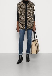 Djurmönstrad väst i beige och svart, matchad med svart polotröja, blå jeans och svarta klackstövlar. Håller en beige axelväska med svarta detaljer.