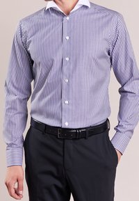 Uomo che indossa una camicia a righe verticali viola e bianche, infilata in pantaloni neri con una cintura nera, con una mano in tasca.