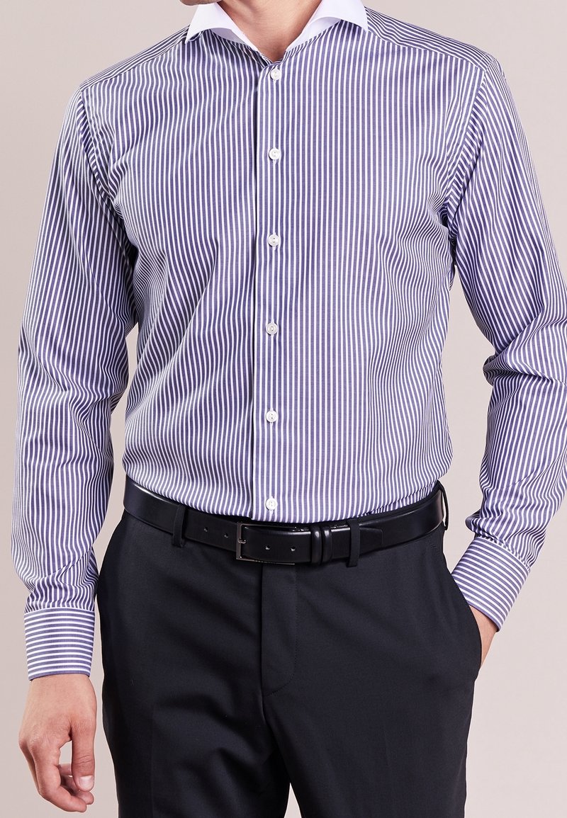 Uomo che indossa una camicia a righe verticali viola e bianche, infilata in pantaloni neri con una cintura nera, con una mano in tasca.