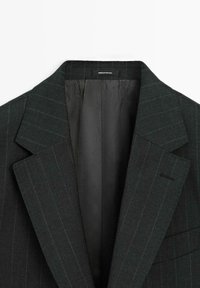 Dunkelgrüner Nadelstreifen-Blazer mit spitzen Revers, einer einzelnen Brusttasche und einem glatten Innenfutter. Das Innenetikett trägt die Aufschrift "Made in Portugal."