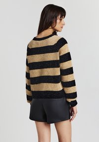 Donna che indossa un maglione a maglia a righe larghe beige e nero e pantaloni in pelle neri, in posizione profilo contro uno sfondo chiaro e semplice.