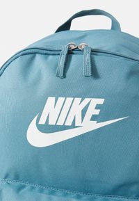 Tirkizni najlon ruksak s dvostrukim zatvaračem, koji se ističe istaknutim bijelim Nike logotipom i čvrstom ručkom na vrhu. Glatka tekstura i lagan dizajn.